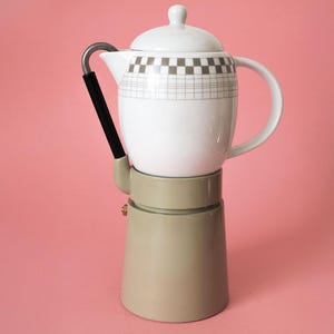 Op de afbeelding: Een koffiezetapparaat met een witte keramische bovenpot met een grijs ruitpatroon en een deksel, bevestigd op een licht olijfgroene metalen voet. Het apparaat heeft een gebogen metalen tuit met een zwart handvat en een zijhandgreep.