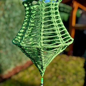 Könnte beinhalten: Eine handgefertigte, grüne Häkellaterne mit rautenförmiger Form. Die Laterne hat ein offenes, netzartiges Design und wird von grünen Schnüren gehalten. Eine Quaste mit Perlen hängt von unten. Der Hintergrund ist verschwommen und zeigt Grün.