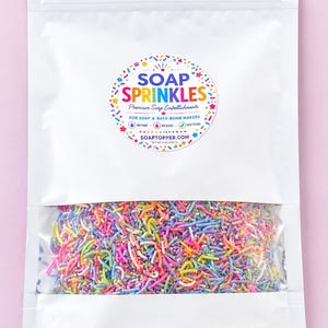 May include: A white stand-up pouch containing multi-colored sprinkles. The label reads SOAP SPRINKLES Premium Soap Embellishments FOR SOAP & BATH BOMB MAKERS NO FADE NO BLEED EASY TO USE SOAPTOPPER.COM NET WT 8 OZ (226 G).