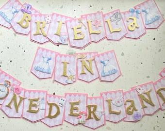 May include: A pink and white banner with gold glitter letters spelling "Briella in Wonderland". The banner features images of Alice in Wonderland characters, including the Mad Hatter, the Cheshire Cat, and the Queen of Hearts.