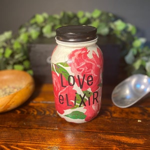 Puede incluir: Un tarro de vidrio blanco con tapa negra decorado con rosas rosas y el texto "Love elixir".