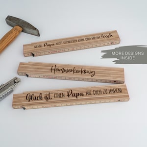 Könnte beinhalten: Drei Holzgliedermaßstäbe mit Gravur, Hammer und Cutter. Die Lineale tragen Sätze wie "Heimwerkerkönig" und "Glück ist einen Papa wie dich zu haben!" in hellem Holz.