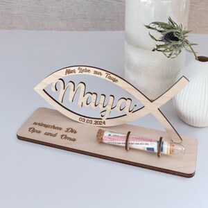 Pode incluir: Um suporte de madeira clara com um recorte em forma de peixe gravado com o nome Maya e a data 03.03.2024. A base inclui o texto Alles Liebe zur Taufe e wunschen Dir Opa und Oma, com um tubo de vidro fixado na estrutura de madeira.