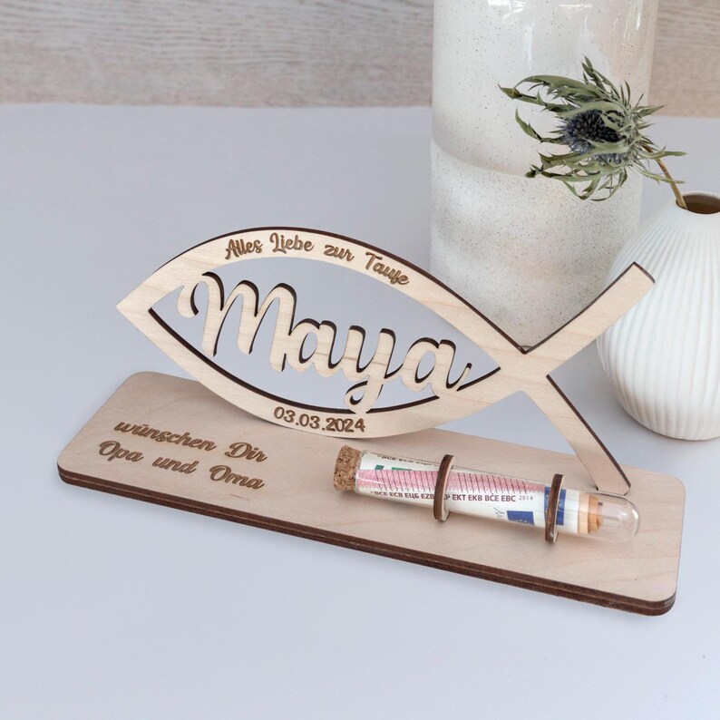Pode incluir: Um suporte de madeira clara com um recorte em forma de peixe gravado com o nome Maya e a data 03.03.2024. A base inclui o texto Alles Liebe zur Taufe e wunschen Dir Opa und Oma, com um tubo de vidro fixado na estrutura de madeira.