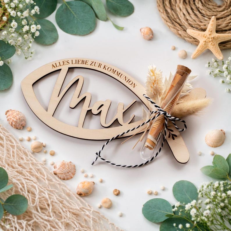 K&ouml;nnte beinhalten: Ein h&ouml;lzernes Taufgeschenk mit dem Namen "Max" und dem Text "Alles Liebe zur Kommunion" eingraviert. Das Geschenk in Fischform ist mit Trockenblumen, einem Reagenzglas und einer schwarz-wei&szlig;en Schnur verziert. Muscheln und Gr&uuml;n umgeben das Geschenk.