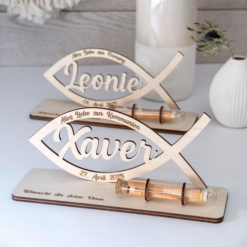 Pode incluir: Dois ornamentos de madeira clara em forma de peixe sobre bases retangulares. Um apresenta o nome Leonie com o texto Alles Liebe zur Firmung e 08. April 2025. O outro apresenta Xaver com Alles Liebe zur Kommunion, 27. April 2025 e W&uuml;nscht dir deine Oma, cada um com um frasco de vidro.
