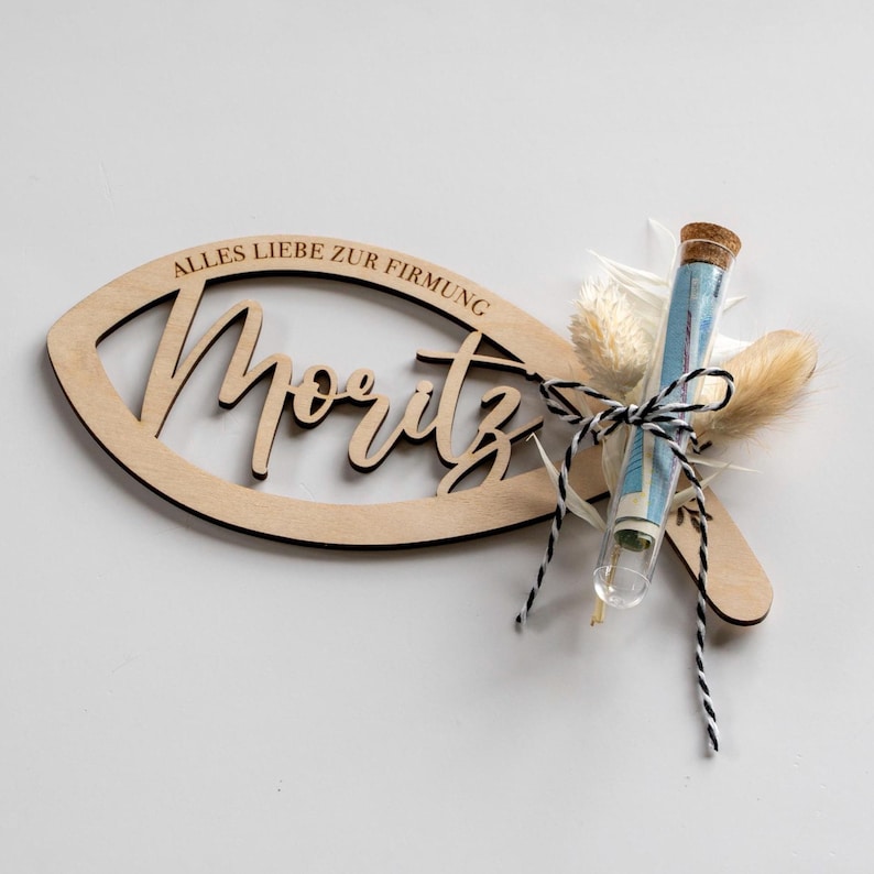 K&ouml;nnte beinhalten: Eine h&ouml;lzerne fischf&ouml;rmige Dekoration mit dem ausgeschnittenen Namen "Moritz" und dem Text "ALLES LIEBE ZUR FIRMUNG". Ein Glasr&ouml;hrchen mit Geld und Trockenblumen ist mit schwarz-wei&szlig;em Bindfaden an der Dekoration befestigt.