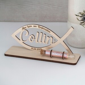 Pode incluir: Uma placa de madeira clara em forma de peixe com o nome Collin, o texto Alles liebe zur Konfirmation e a data 11.05.2025. A placa est&aacute; montada numa base retangular de madeira com um pequeno frasco de vidro contendo fio cor-de-rosa.