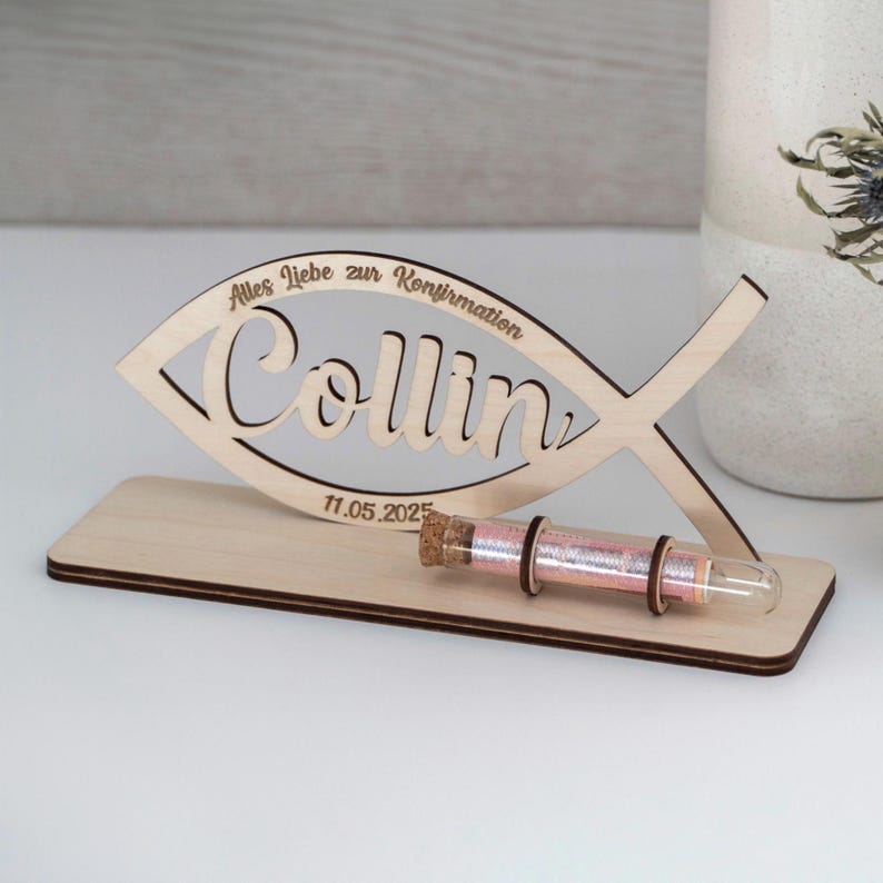 Pode incluir: Uma placa de madeira clara em forma de peixe com o nome Collin, o texto Alles liebe zur Konfirmation e a data 11.05.2025. A placa est&aacute; montada numa base retangular de madeira com um pequeno frasco de vidro contendo fio cor-de-rosa.