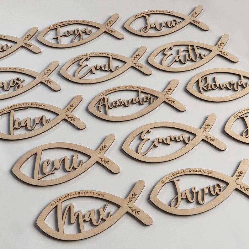 K&ouml;nnte beinhalten: Holzfischf&ouml;rmige Namensschilder mit gelaserten Namen und dem Satz "Alles Liebe zur Konfirmation". Die Namen sind Feya, Jona, Emil, Edith, Ivan, Alexander, Emma, Teni, Max und Jarro. Jedes Schild hat dekorative Bl&auml;tter.