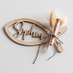 K&ouml;nnte beinhalten: Eine h&ouml;lzerne fischf&ouml;rmige Dekoration mit dem Namen "Sophia" und dem Text "ALLES LIEBE ZUR KONFIRMATION" eingraviert. Ein Reagenzglas mit Geld und Trockenblumen ist mit schwarz-wei&szlig;em Garn an der Dekoration befestigt. Die Dekoration ist aus hellem Holz gefertigt.