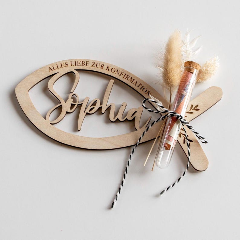 K&ouml;nnte beinhalten: Eine h&ouml;lzerne fischf&ouml;rmige Dekoration mit dem Namen "Sophia" und dem Text "ALLES LIEBE ZUR KONFIRMATION" eingraviert. Ein Reagenzglas mit Geld und Trockenblumen ist mit schwarz-wei&szlig;em Garn an der Dekoration befestigt. Die Dekoration ist aus hellem Holz gefertigt.