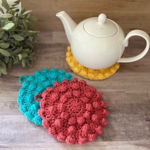 May include: Three crocheted pot holders in teal, red and yellow. The teal and red pot holders are round and have a textured, bumpy surface. The yellow pot holder is under a white teapot.