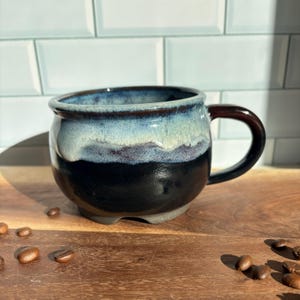 May include: A round ceramic mug featuring a glossy glaze that transitions from a deep black base to a light blue and white rim. The mug has a dark brown handle and a wide, curved body, accompanied by scattered roasted coffee beans.