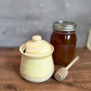 May include: A ceramic honey pot with a lid, a wooden honey dipper, and a glass jar of honey. The honey pot is pale yellow with a white base. The honey dipper is light brown. The jar has a silver lid.
