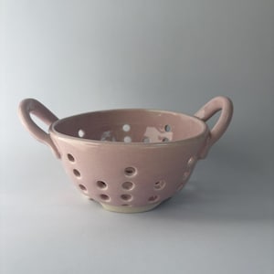 May include: A pink ceramic colander with two curved handles. The bowl has numerous small holes throughout the sides and bottom. The colander is a light pink color and sits on a white base.