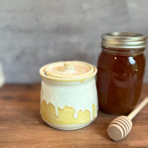 May include: A ceramic honey pot with a lid, featuring a white and yellow glaze. A glass jar of honey and a wooden honey dipper are next to the pot. The honey pot has a dripped glaze design.