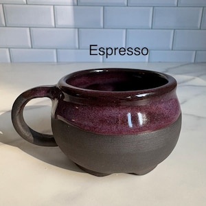 May include: A ceramic mug with a rounded shape and a single handle. The upper portion features a glossy, deep purple glaze, while the lower portion has a matte, dark charcoal finish. The word Espresso is printed in black above the mug.