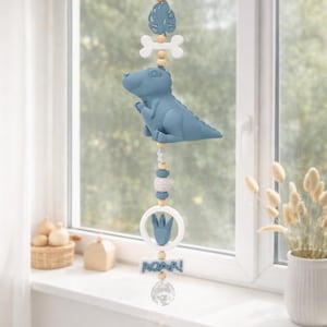 Könnte beinhalten: Ein Mobile zum Thema Dinosaurier in Blau-, Weiß- und Holztönen. Es zeigt einen blauen Dinosaurier, einen Knochen, ein Blatt und ein Handdesign, mit dem Wort "ROAR!" am unteren Rand. Das Mobile ist an einer Schnur aufgehängt.