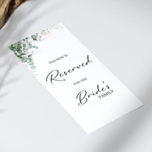 Op de afbeelding: Een wit rechthoekig bord met de tekst "THIS ROW IS Reserved FOR THE Bride's FAMILY" in elegant zwart schrift. De linkerbovenhoek heeft een aquarelontwerp van groene en gouden bladeren.