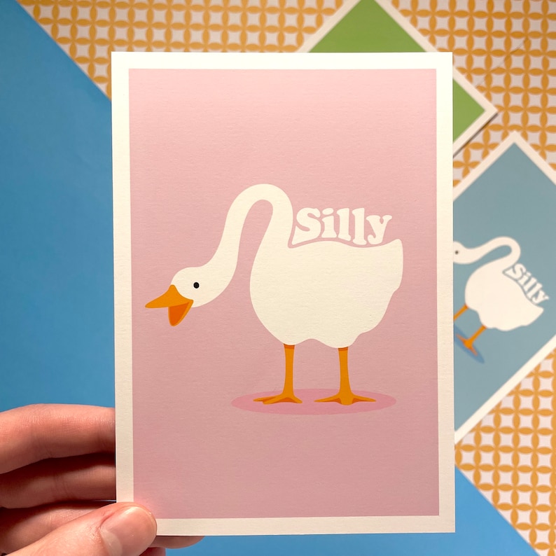 K&ouml;nnte beinhalten: Eine rosa Karte mit einer Illustration einer wei&szlig;en Gans. Die Gans hat den Kopf nach hinten gedreht und das Wort "Silly" steht auf ihrem R&uuml;cken.