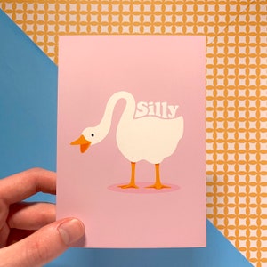 K&ouml;nnte beinhalten: Eine rosa Karte mit einer Illustration einer wei&szlig;en Gans. Die Gans hat den Kopf nach links gedreht und den Hals nach hinten gebogen. Das Wort "Silly" ist in wei&szlig;en Buchstaben auf dem R&uuml;cken der Gans geschrieben.