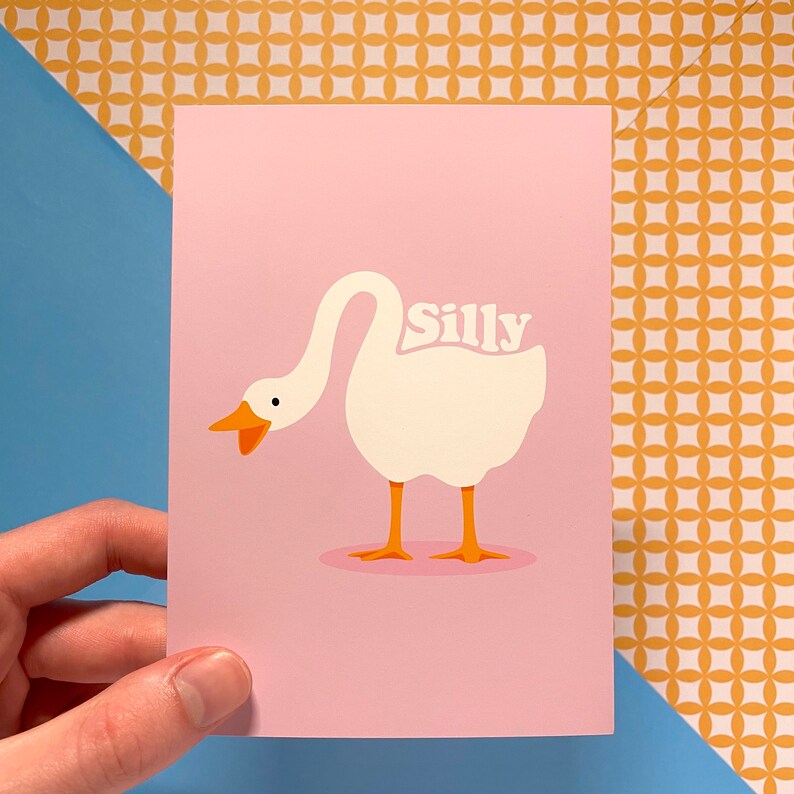 K&ouml;nnte beinhalten: Eine rosa Karte mit einer Illustration einer wei&szlig;en Gans. Die Gans hat den Kopf nach links gedreht und den Hals nach hinten gebogen. Das Wort "Silly" ist in wei&szlig;en Buchstaben auf dem R&uuml;cken der Gans geschrieben.