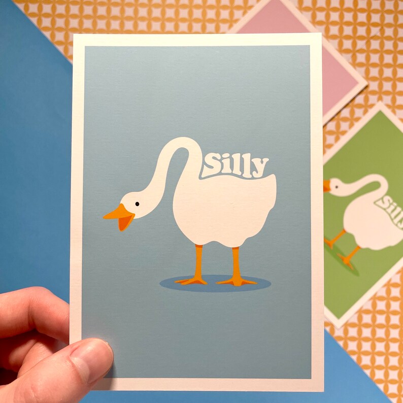 K&ouml;nnte beinhalten: Eine hellblaue Karte mit einer Illustration einer wei&szlig;en Gans. Die Gans schaut nach links und hat das Wort "silly" in einer Retro-Schrift auf ihrem R&uuml;cken geschrieben.