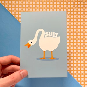 K&ouml;nnte beinhalten: Eine hellblaue Karte mit einer Illustration einer wei&szlig;en Gans. Die Gans hat einen orangefarbenen Schnabel und F&uuml;&szlig;e. Das Wort "Silly" ist in wei&szlig;en Buchstaben auf dem R&uuml;cken der Gans geschrieben.