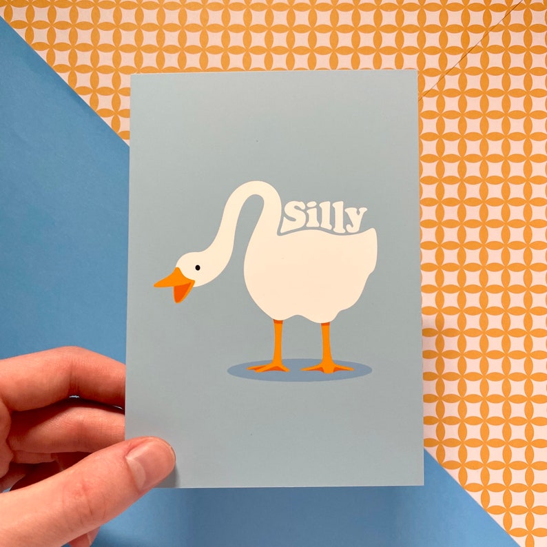K&ouml;nnte beinhalten: Eine hellblaue Karte mit einer Illustration einer wei&szlig;en Gans. Die Gans hat einen orangefarbenen Schnabel und F&uuml;&szlig;e. Das Wort "Silly" ist in wei&szlig;en Buchstaben auf dem R&uuml;cken der Gans geschrieben.