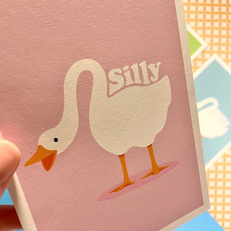 K&ouml;nnte beinhalten: Eine rosa Karte mit einer Illustration einer wei&szlig;en Gans. Die Gans schaut nach unten, den Kopf nach hinten gebeugt. Das Wort "silly" ist in einer Retro-Schrift auf dem R&uuml;cken der Gans geschrieben.