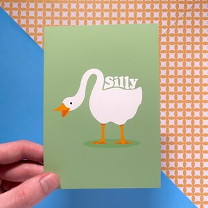 K&ouml;nnte beinhalten: Eine gr&uuml;ne Karte mit einer Illustration einer wei&szlig;en Gans. Die Gans hat den Kopf zur&uuml;ckgedreht und das Wort "Silly" steht auf ihrem R&uuml;cken.
