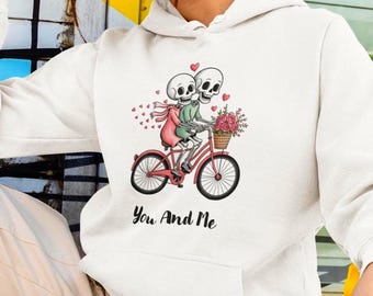 May include: White hoodie featuring a graphic of two skeleton figures riding a red bicycle, with a basket of flowers. The phrase "You And Me" is printed below the bicycle. Hearts and a pink scarf add detail.