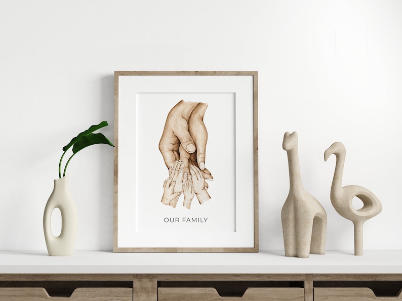Op de afbeelding: Een ingelijste aquarel illustratie van handen die elkaar vasthouden. De handen zijn bruin en de achtergrond is wit. De tekst "OUR FAMILY" is onder de handen gedrukt.