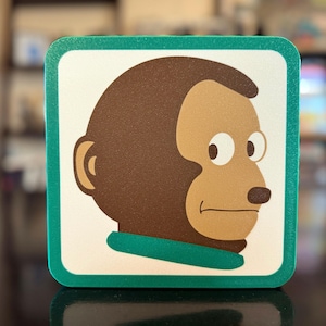 May include: Square coaster featuring a cartoon monkey with a brown head and tan face. The monkey has a green collar and is set against a white background with a green border. The coaster is likely made of a durable material.