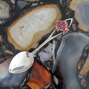 May include: A silver spoon with a red and blue fish design on the handle. The spoon is lying on a surface made of many different colored agate stones.