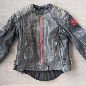 May include: A distressed gray leather motorcycle jacket with a red zipper and accents. The jacket has a round collar, zippered pockets, and a red emblem on the upper arm. The jacket is laid flat on a wooden surface.