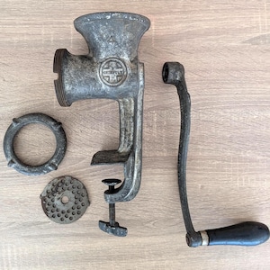 May include: An antique, cast-iron meat grinder with a clamp, handle, and several detachable parts. The grinder is a dull silver color, with a black handle. The parts are arranged on a light wood surface.