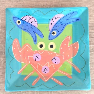 Puede incluir: Plato cuadrado de cerámica con un tema oceánico pintado a mano. El plato presenta un cangrejo en el centro, dos peces azules y un borde turquesa. El cangrejo es naranja con detalles morados, y los peces son azules con detalles rosas.