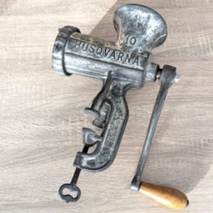 May include: Vintage Husqvarna meat grinder, a cast-iron kitchen tool with a hand crank and wooden handle. The grinder is silver-toned with the brand name embossed on the side. It has a clamp for securing it to a surface.