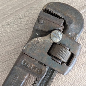 May include: Close-up of a vintage TRIMO 12-inch pipe wrench. The wrench is made of dark metal with visible wear and tear. The jaws are serrated, and the handle features the brand name. The adjustable jaw is controlled by a knurled wheel.