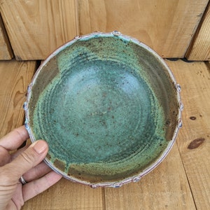 May include: A green and brown ceramic bowl with a textured rim. The bowl has a speckled glaze and a slightly uneven surface.