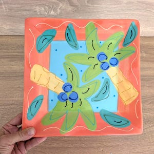 Puede incluir: Plato cuadrado de cerámica con un diseño tropical pintado a mano. El plato presenta un borde de color coral con un cuadrado azul claro en el centro. Dos palmeras verdes con cocos azules están pintadas en el plato, junto con hojas turquesas y troncos amarillos.