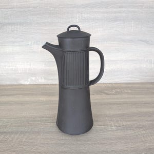 Puede incluir: Una cafetera negra mate con tapa y asa. La cafetera tiene un diseño acanalado alrededor del centro y un pico para verter. La cafetera está sobre una superficie de madera.