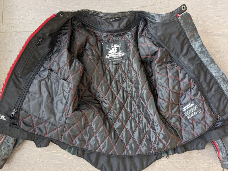 May include: Black leather motorcycle jacket with red accents. The interior features a quilted black lining with red stitching. The jacket has multiple pockets and a zipper closure. The brand logo is visible on the inside.