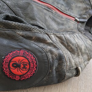 May include: Black leather motorcycle jacket with a distressed finish. Features a red zipper and a round patch with a spider graphic and the words "Speed and Strength". Perforated panels and quilted stitching are visible.