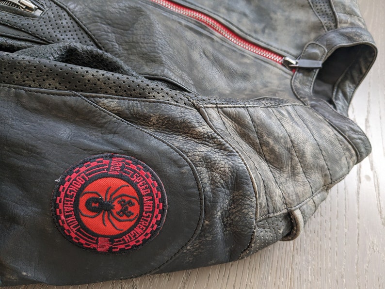 May include: Black leather motorcycle jacket with a distressed finish. Features a red zipper and a round patch with a spider graphic and the words "Speed and Strength". Perforated panels and quilted stitching are visible.