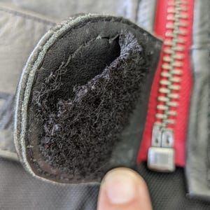 May include: Close-up of a black leather jacket detail. The image shows a worn, rounded patch with a frayed interior, and a red zipper is visible in the background.