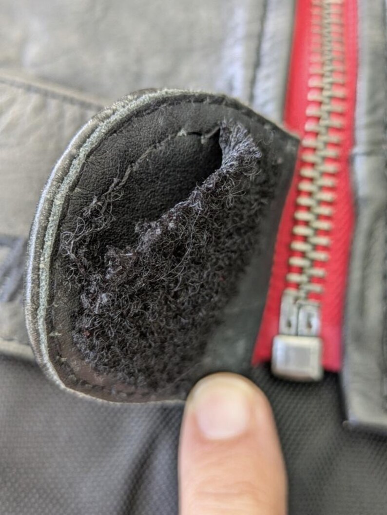 May include: Close-up of a black leather jacket detail. The image shows a worn, rounded patch with a frayed interior, and a red zipper is visible in the background.