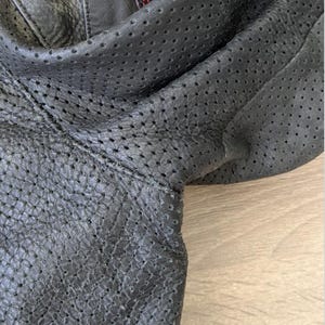 May include: Close-up of a black leather jacket with perforated detailing. The jacket has a patch with red and white text. The leather appears textured and the jacket is folded.